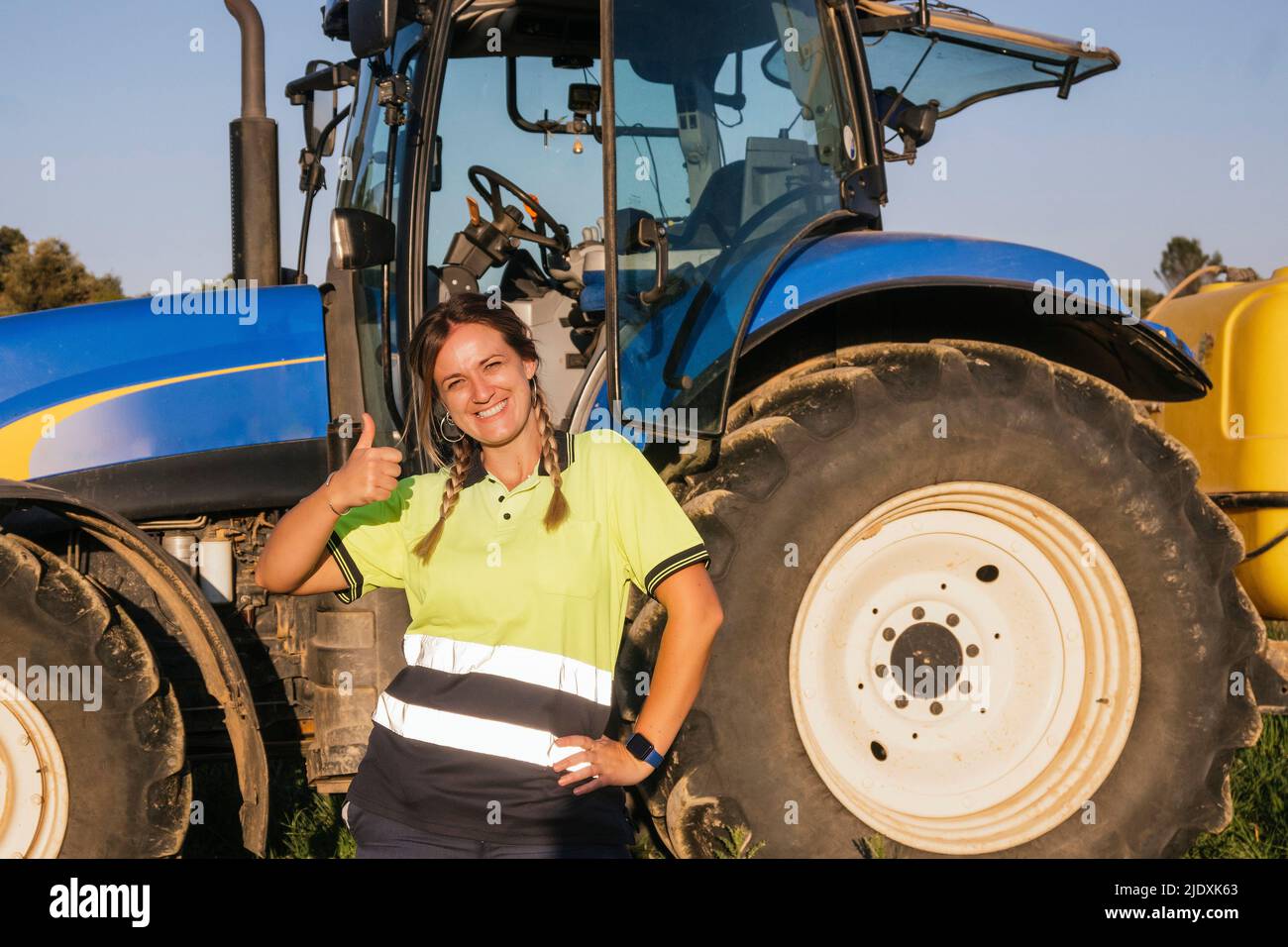 Happy female farmer gesturing thumbs up in front of tractor on sunny ...