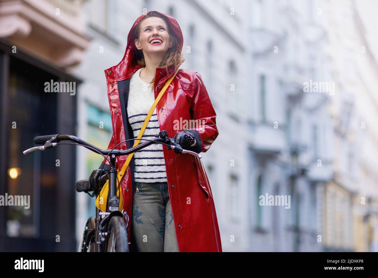 smiling modern 40 years old woman in red rain coat with bicycle in the ...