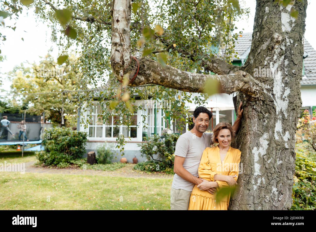 Happy couple standing by tree in front of house Stock Photo - Alamy