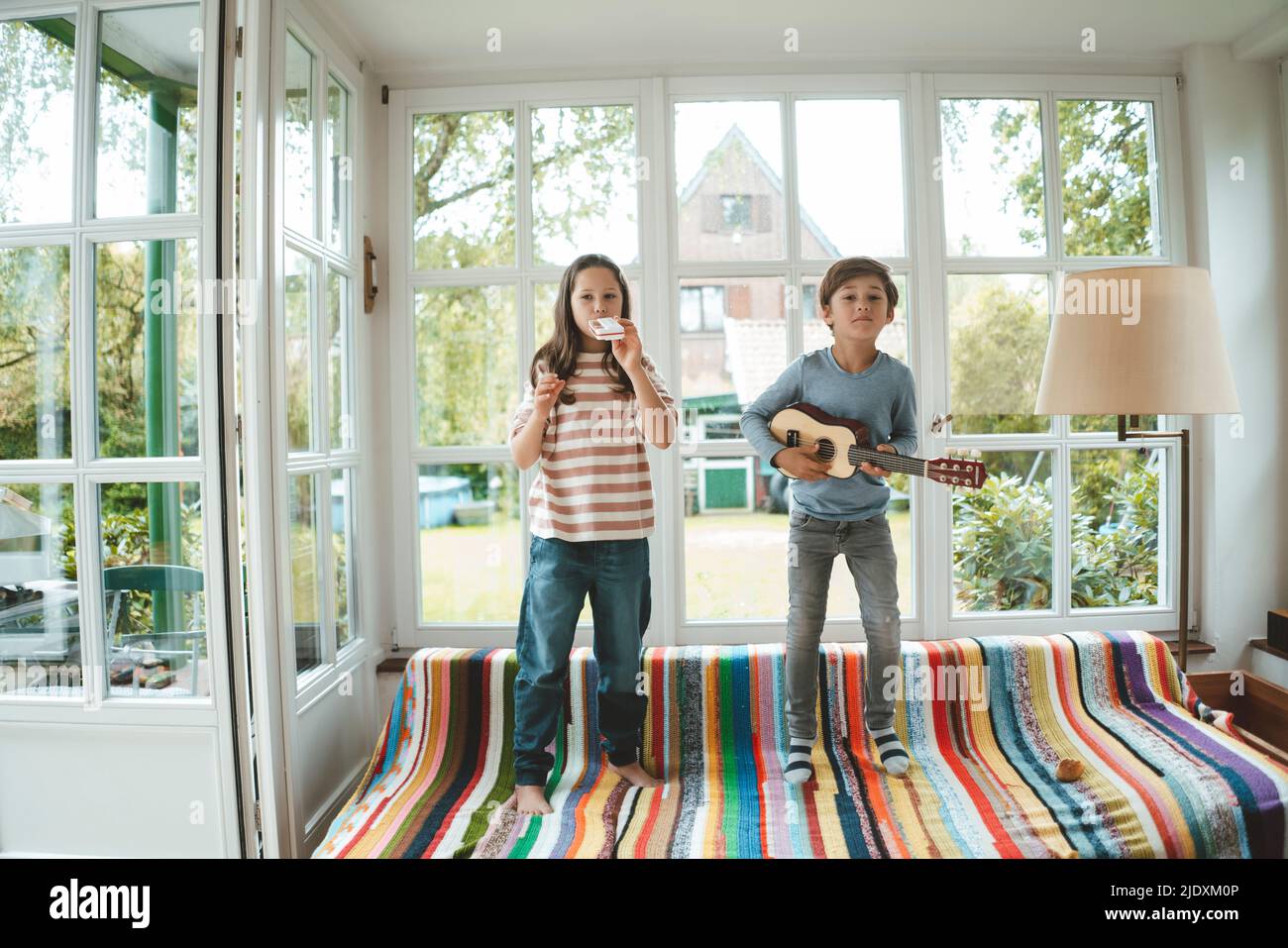 Siblings playing musical instrument standing on sofa at home Stock Photo
