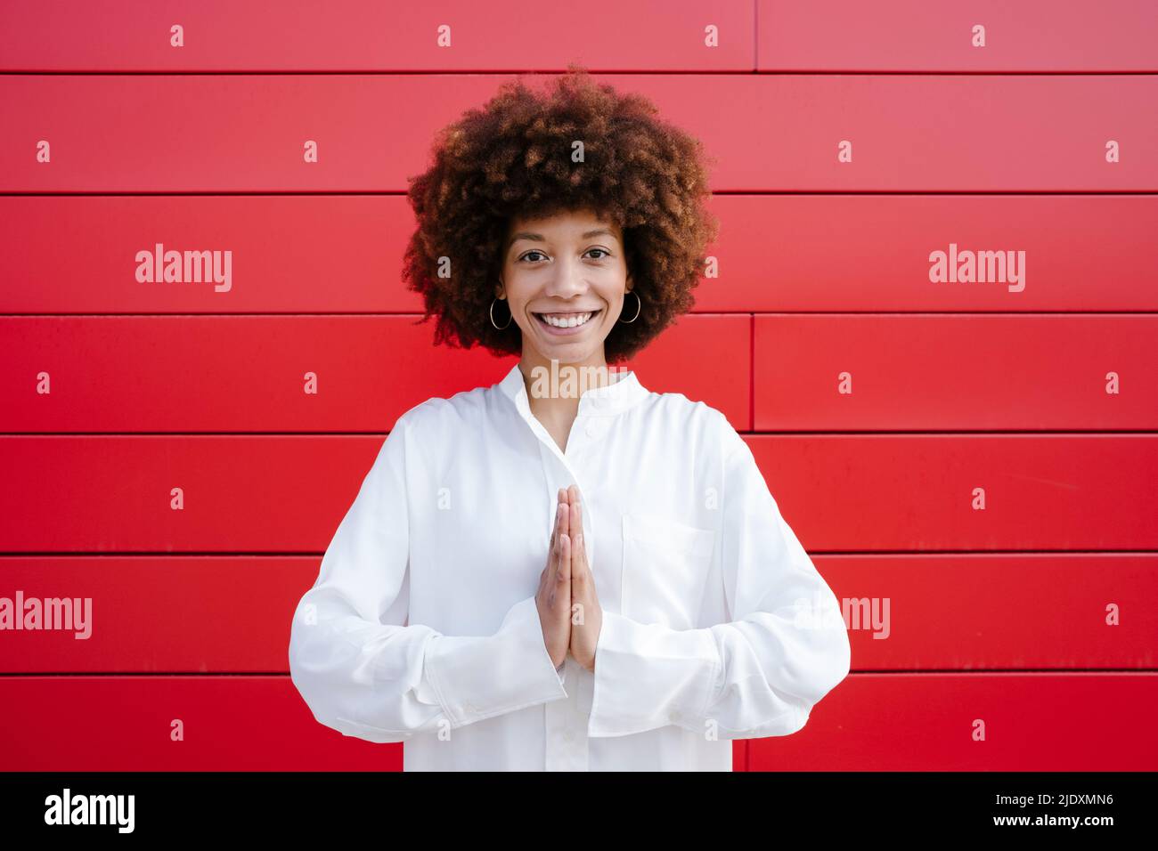 Smiling Afro woman standing with hands clasped in front of red wall ...