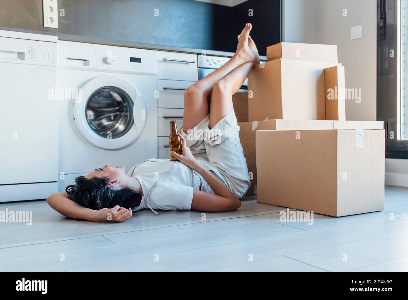 Happy woman holding beer bottle with feet up on cardboard box lying by washing machine in