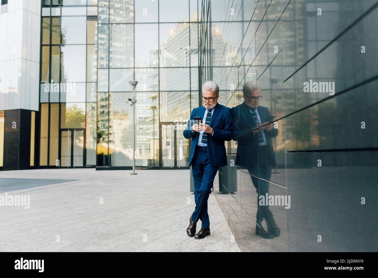 Senior businessman using smart phone standing with legs crossed at ankle outside office building Stock Photo