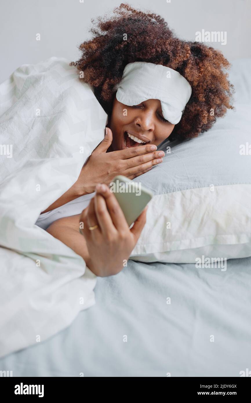 African woman yawns in bed and looks at messages on the phone Stock Photo