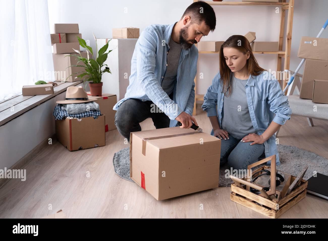 A man and a woman on the day of the move sit on the floor and unpack things. A man cuts the adhesive tape on a cardboard box with a knife. Happy coupl Stock Photo