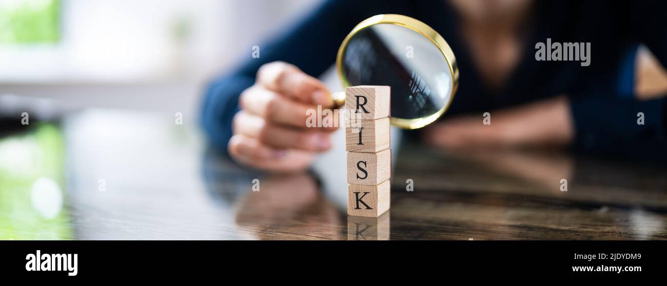 Close-up Of Wooden Blocks With Risk Word In Front Of Businessperson's Hand Holding Magnifying Glass Stock Photo