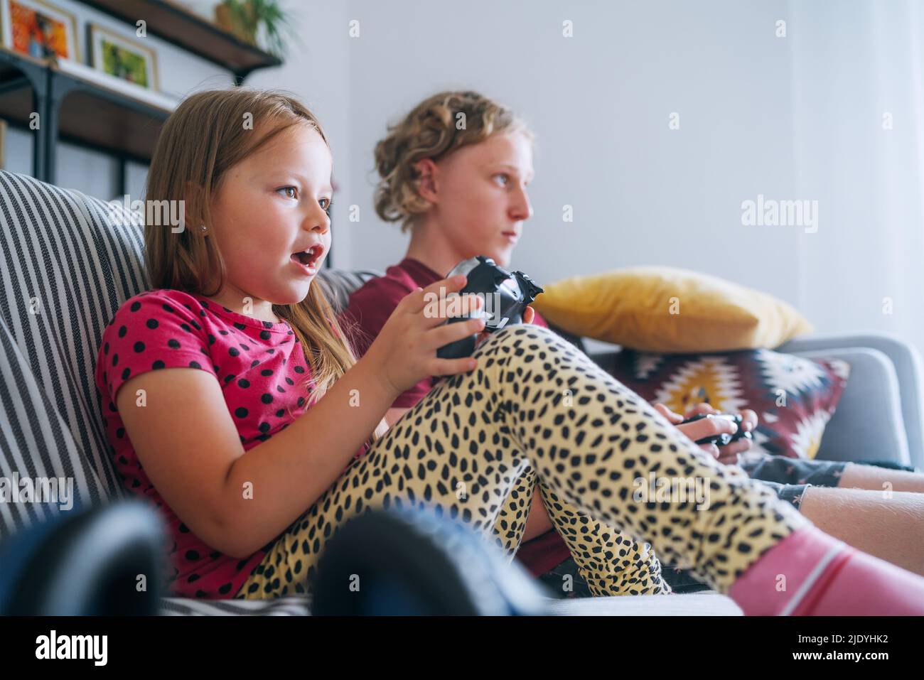 Smiling little Sister with brother sitting on sofa in living room, looking TV and playing console video game using wireless controllers. Careless chil Stock Photo