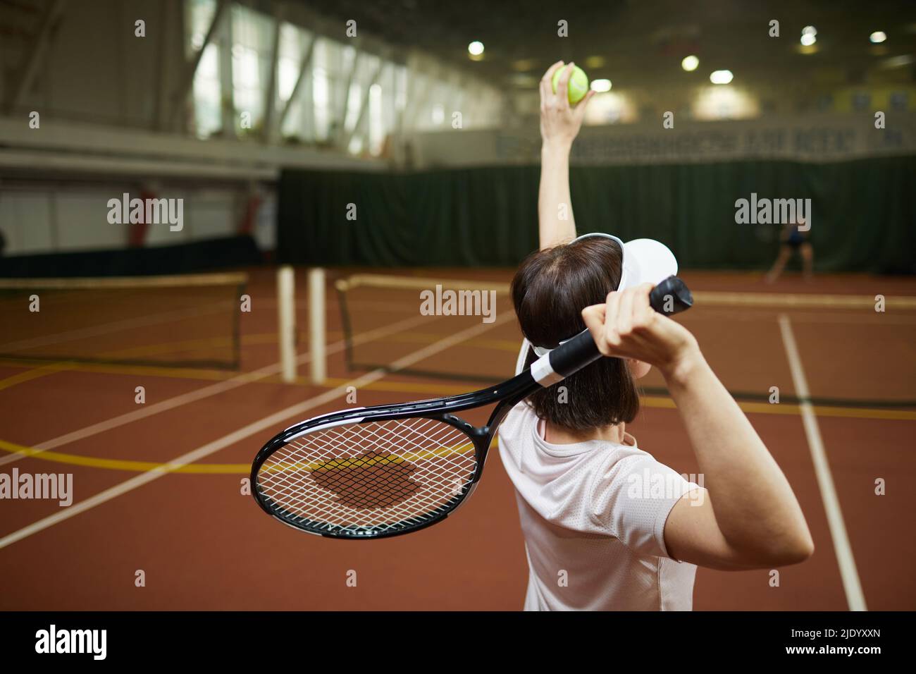Rear view of forceful brunette woman in cap standing on tennis court and swinging arm with racket for hitting ball Stock Photo