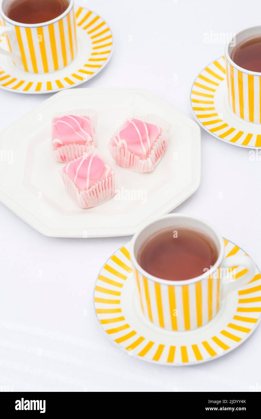 Afternoon tea. Black tea served in bright yellow striped cups and saucers with pink fondant coated sponge cakes. Stock Photo