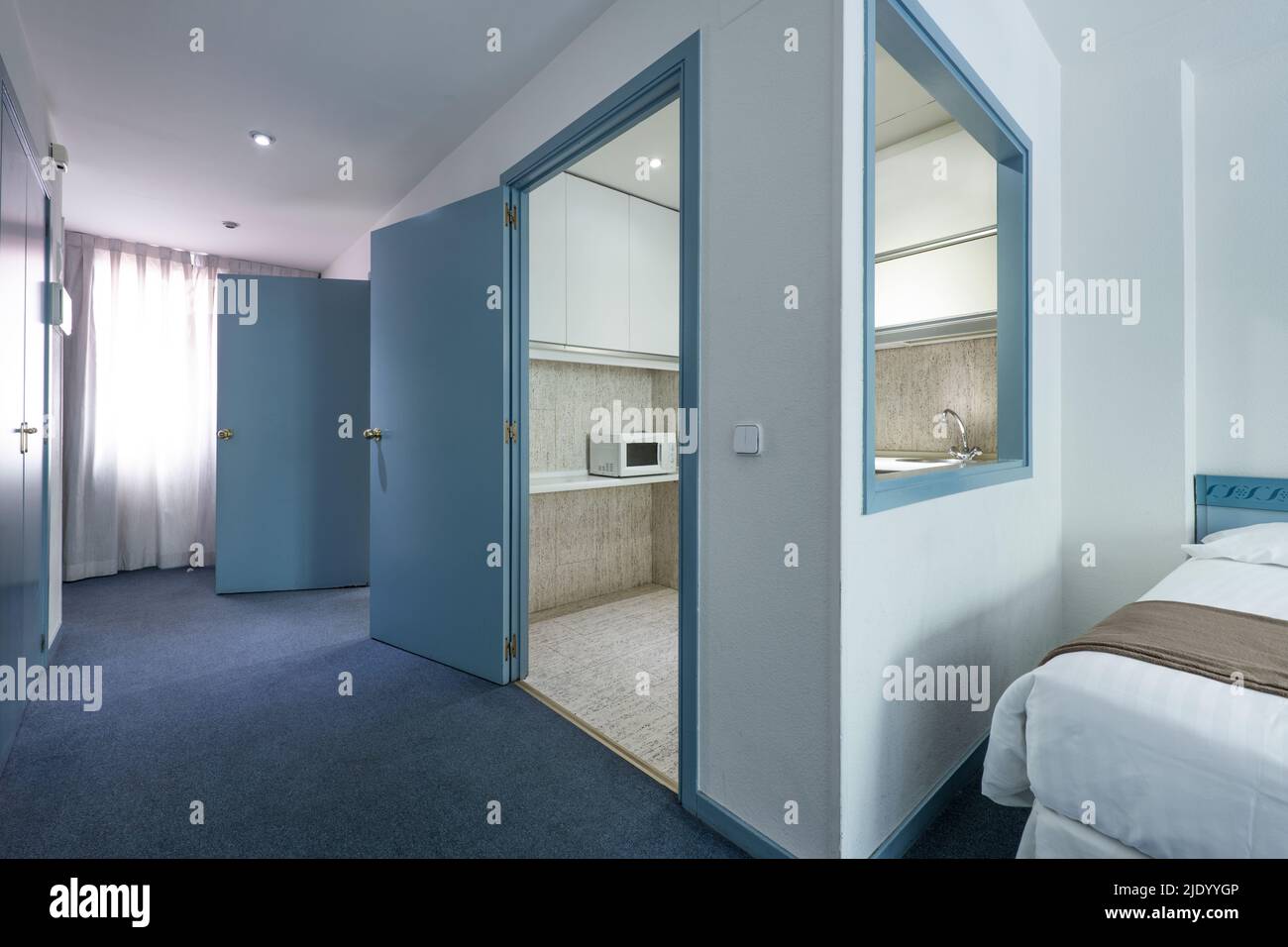 Cheap hotel room with blue carpet floors, serving hatch in the kitchen, and brown blankets on white duvets Stock Photo