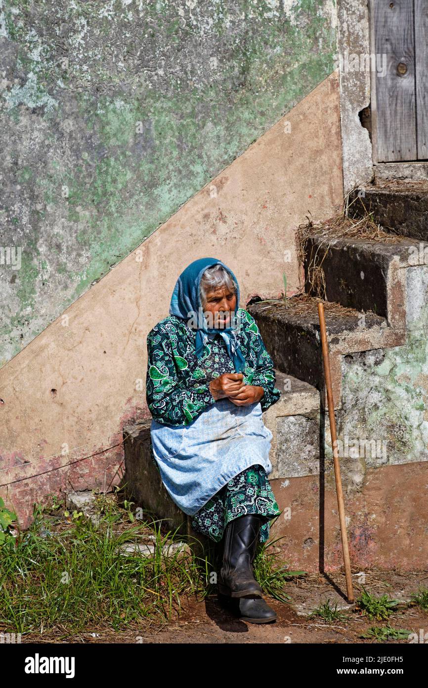 Old woman, peasant with headscarf, apron, boots and stick sitting on stairs in front of stone house, Faial, Madeira, officially Autonomous Region of Stock Photo