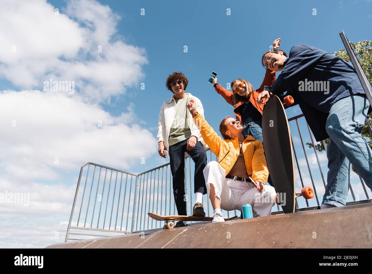 low angle view of cheerful multiethnic friends on skate ramp against blue cloudy sky Stock Photo