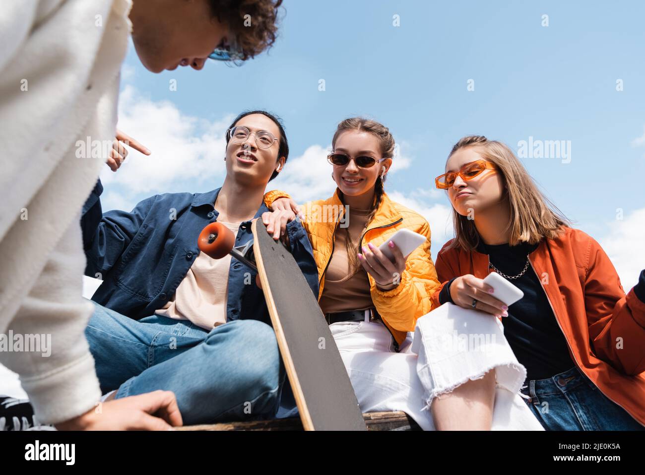 low angle view of asian skater pointing with finger near friends with smartphones outdoors Stock Photo