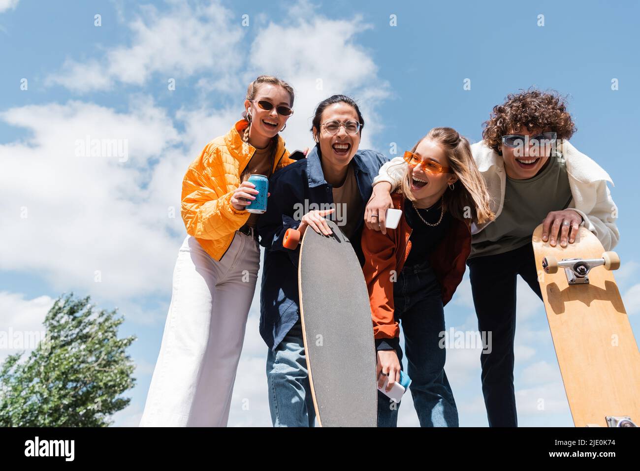 low angle view of interracial skaters near laughing friends with smartphones and soda cans against cloudy sky Stock Photo