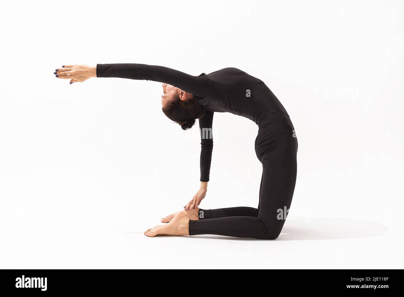 A woman in a black tracksuit practicing yoga performs a variation of the Ushtrasana exercise, camel pose with outstretched arm, on a white background Stock Photo