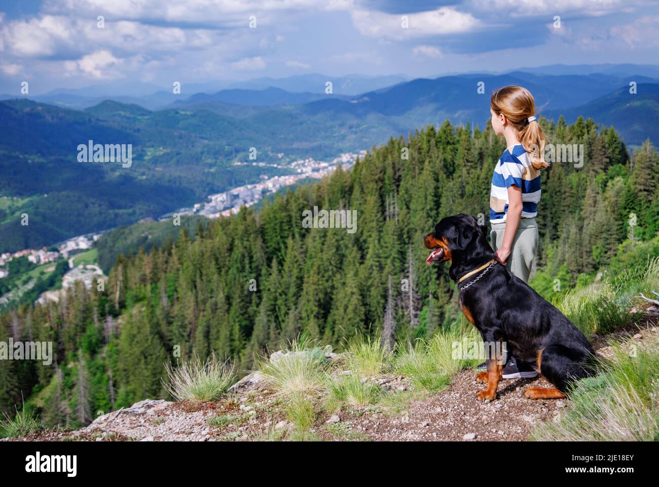 A little pensive dreamy girl with blond hair stands next to her big obedient faithful dog of the Rottweiler breed on a high rocky peak with mountain v Stock Photo