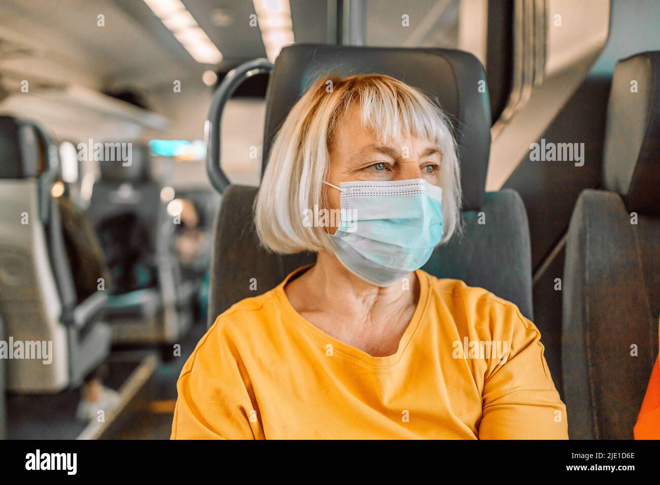 Panoramic banner of 50s blonde woman wearing protective face mask on train looking through the window.Travel safely on public transport.  Stock Photo