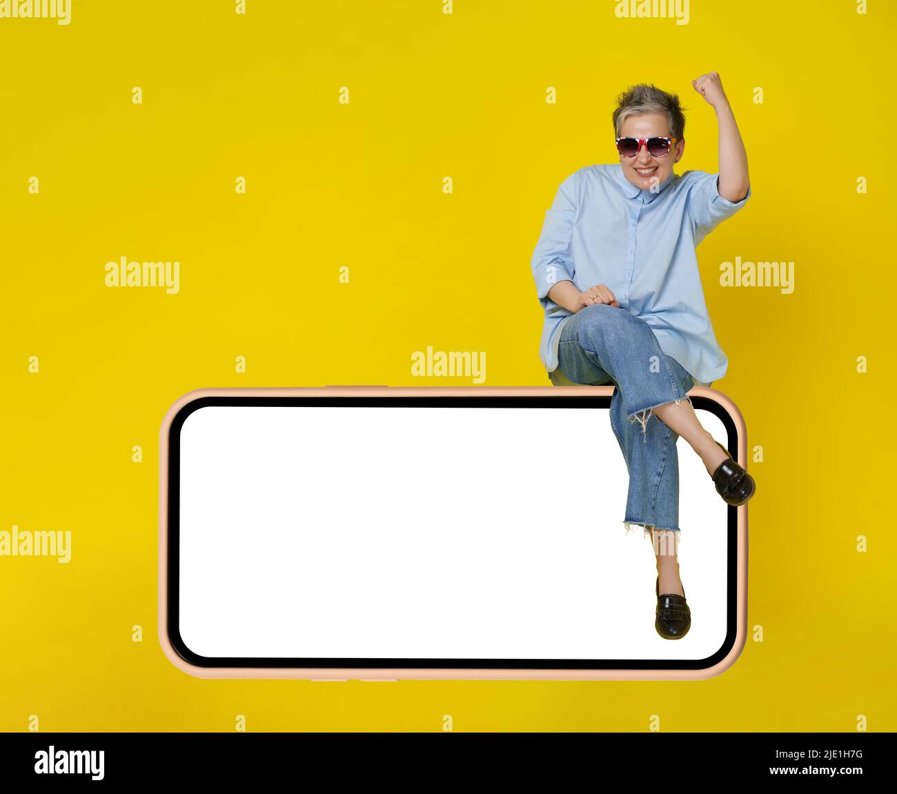 Crazy mad mature grey haired woman sitting on a giant, huge smartphone in casual blue shirt and jeans, sunglasses and hands lift up in a win, isolated on yellow background.  Stock Photo