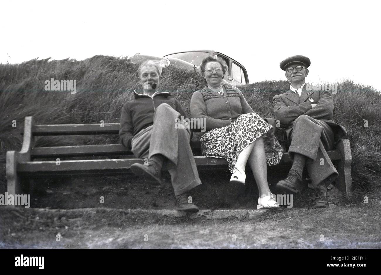 1940s, historical, a day out, sitting on a bench at the coast, England, UK, the men on the left of the lady is smoking a pipe, while the gentleman on right with the moustache is wearing a cloth cap and his army medals on his jacket. Stock Photo