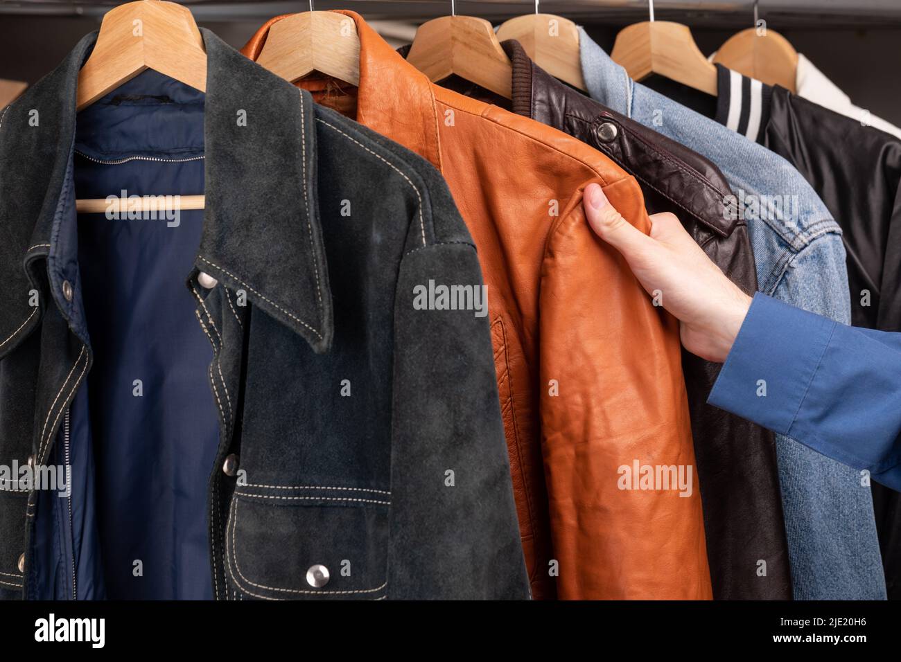 Man choosing clothing in a second hand store. Various vintage suede leather and jeans jackets hang on clothing rack. Thrifting and sustainability in c Stock Photo