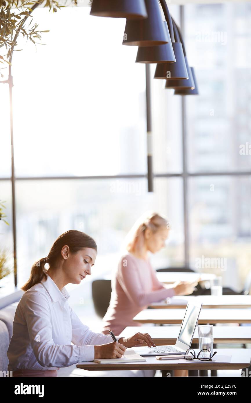 Serious beautiful young businesswoman in formal outfit sitting at table in cafe and making notes in diary while planning her day Stock Photo