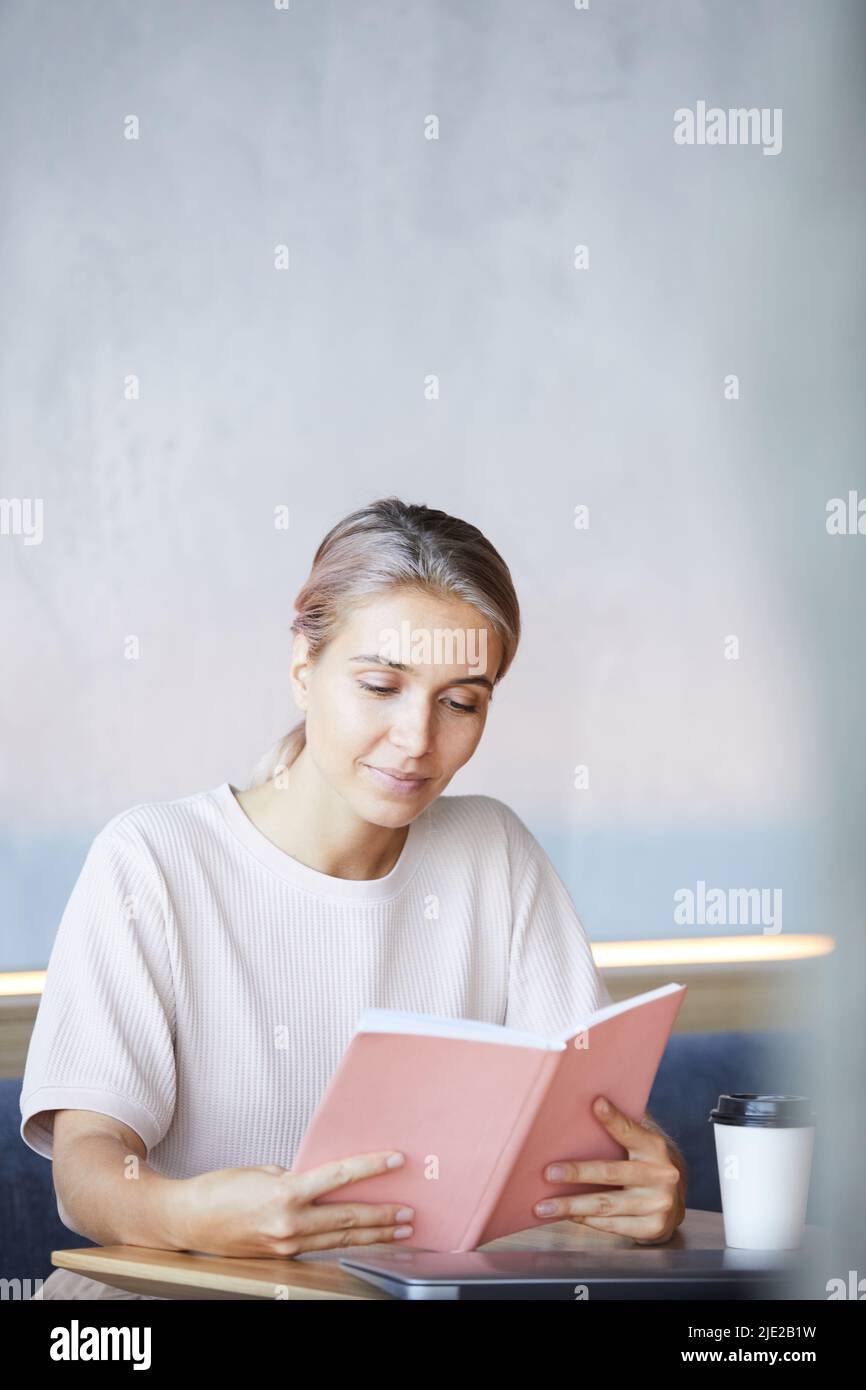 Content curious young lady sitting at table with coffee cup and spending time with interesting book Stock Photo