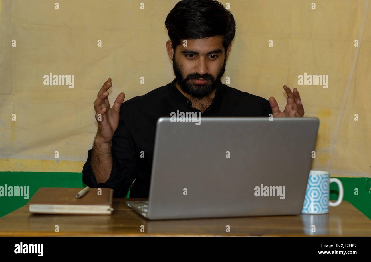 A young Indian man sitting on a chair and giving angry expression looking at laptop screen. Diary and coffee mug on table. Selective focus on face. Stock Photo