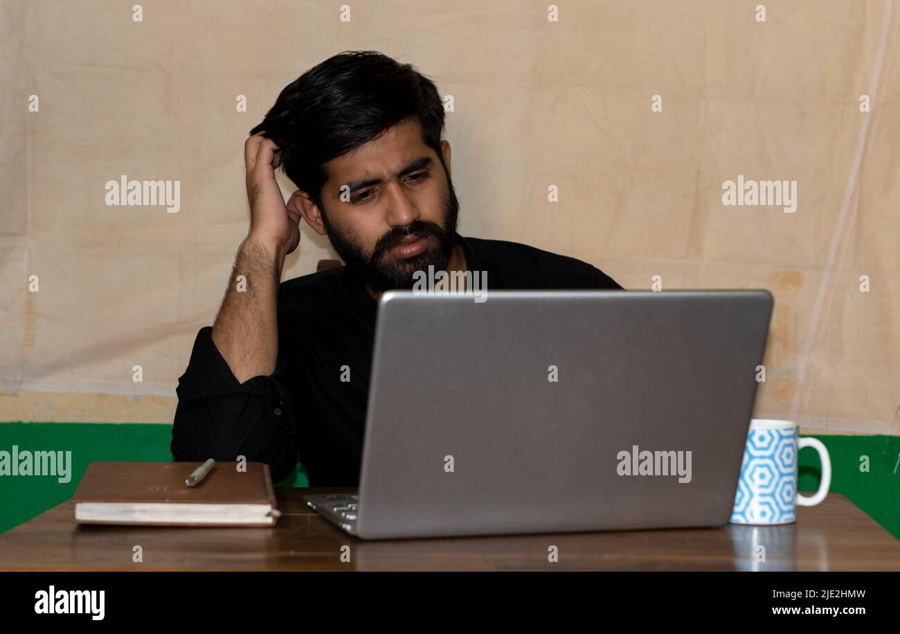 A young Indian man sitting on a chair and giving confused expression looking at laptop screen. Diary and coffee mug on table. Selective focus on face. Stock Photo
