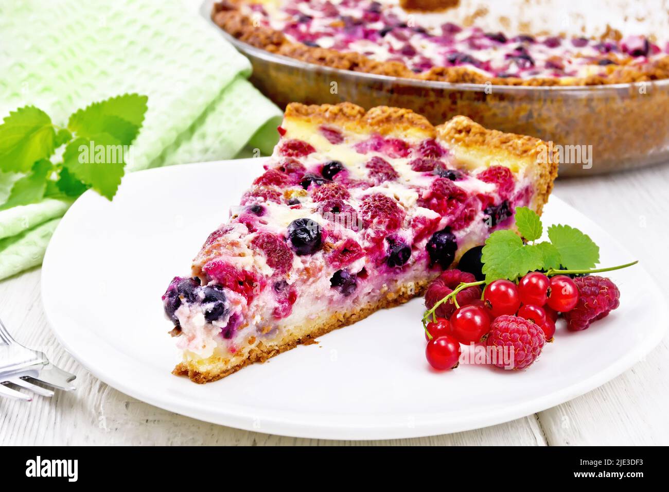 Pie with raspberries, currants and cream sauce in a plate, towel and mint on wooden board background Stock Photo