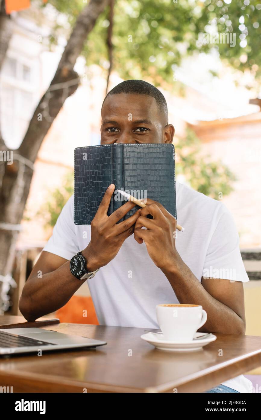 Vertical smiling, glad african american man sitting at the table with laptop covering his mouth by book in coffee shop Stock Photo