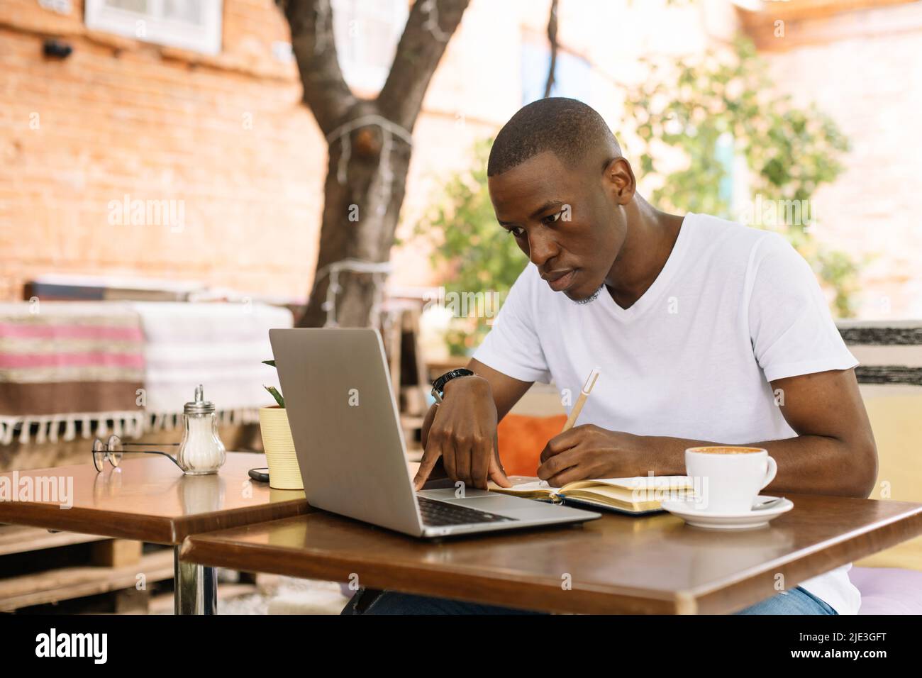 Serious, concentrated and enthusiastic multiracial man using laptop, writing notes in workbook in cafe. Student and exam Stock Photo