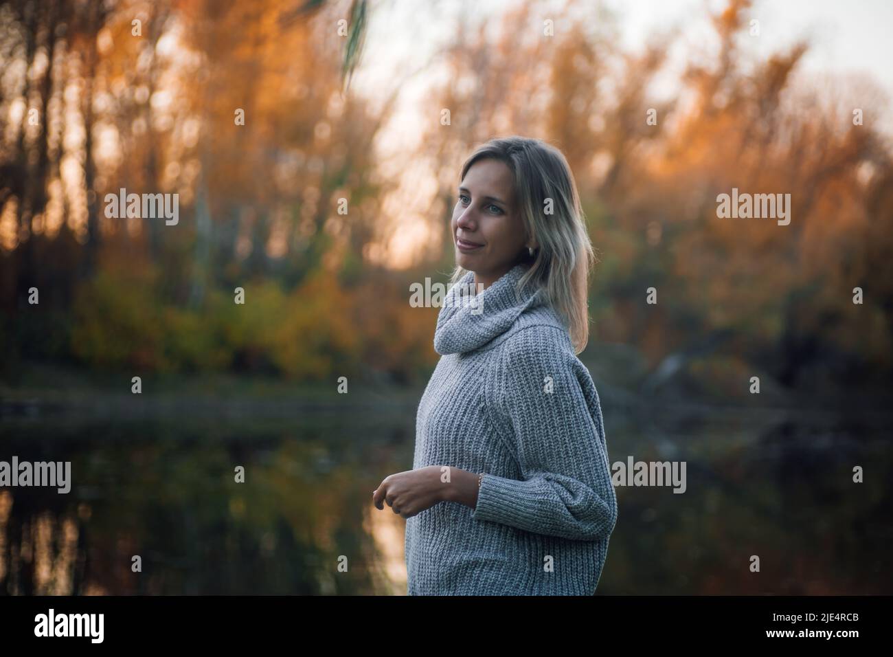 Beautiful blonde woman standing by river in forest, looking away smiling and posing with trees covered with sunset in background, wearing grey sweater Stock Photo