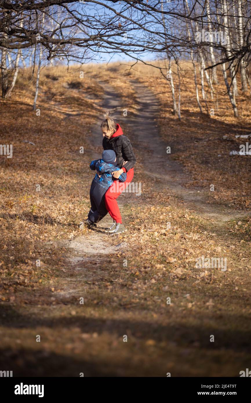 Happy mother playing with young son both smiling having walk on country road in forest with dry leaves lying on ground on sunny autumn day. Family Stock Photo