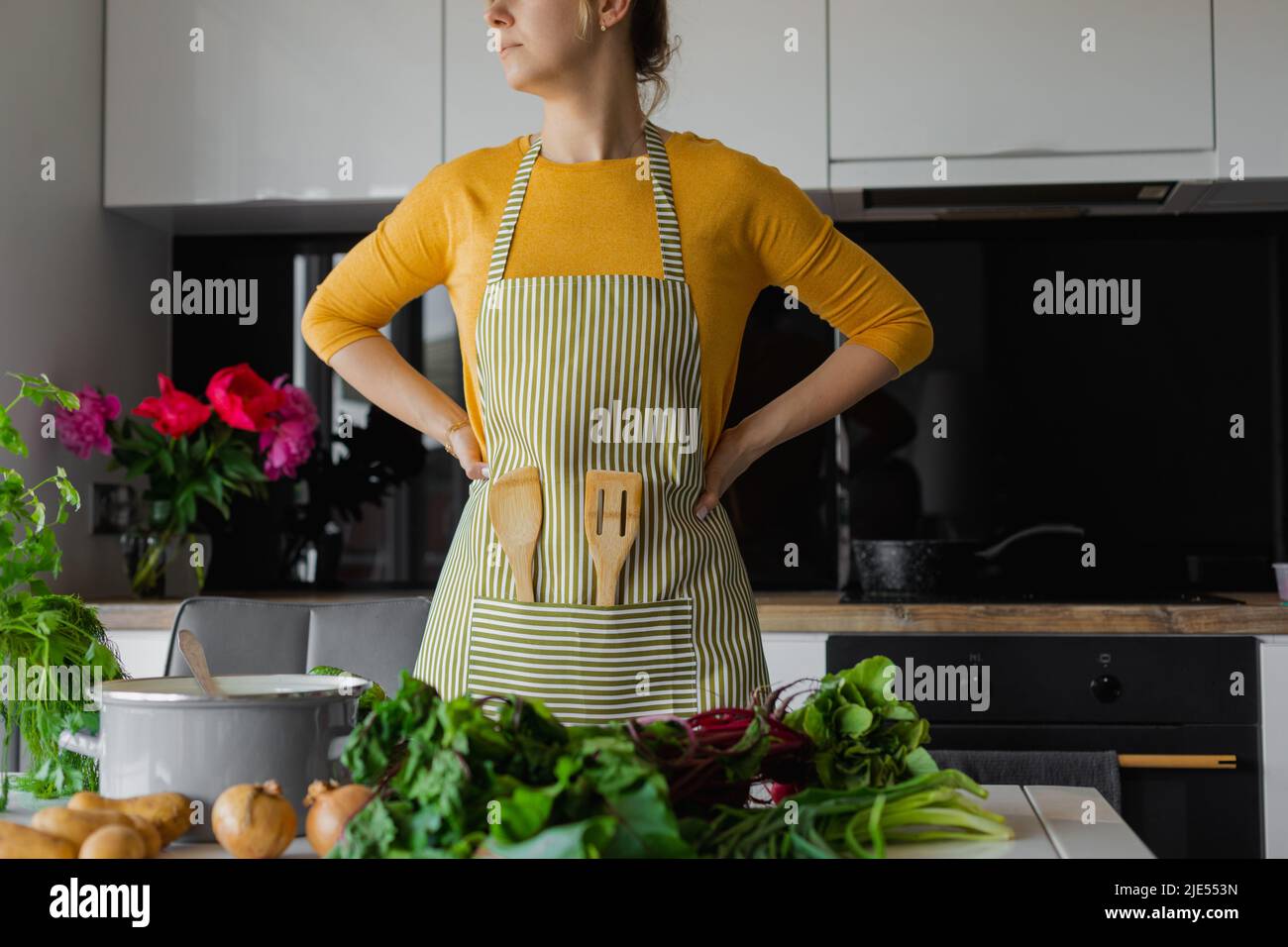 Cropped blonde woman in striped kitchen apron with wooden spatula utensil inside. Female arms akimbo, choose ingredients Stock Photo