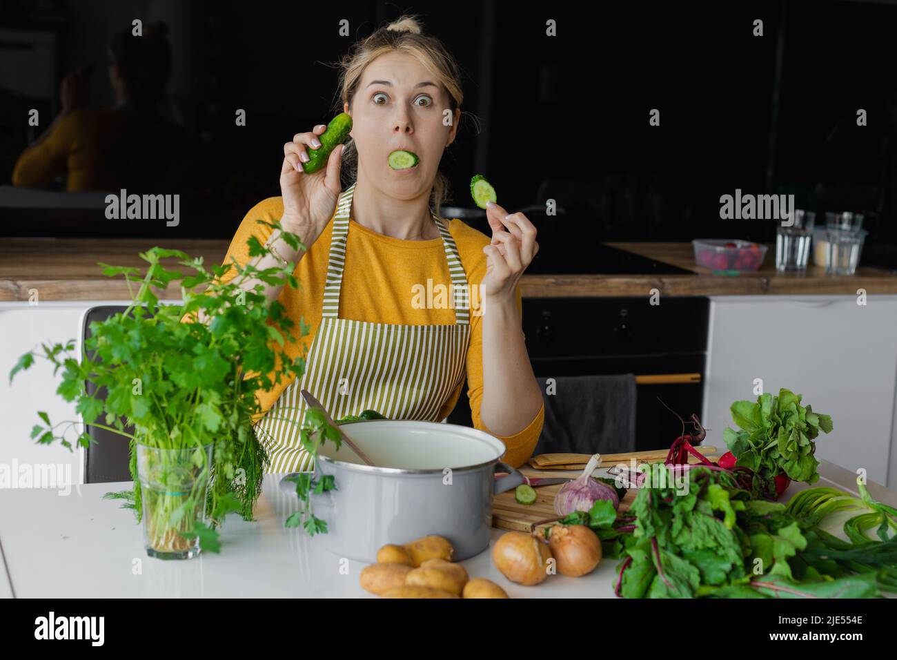 Funny and surprised blond woman with cucumber in mouth, cooking fresh mixed vegetables for salad. Summer organic recipe Stock Photo