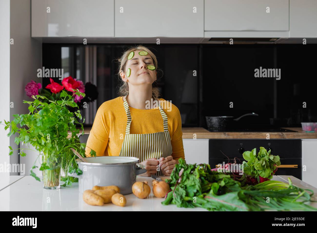 Funny relaxed blond woman with sliced circle cucumber on face, mask in kitchen. Fresh vegetables for skin care treatment Stock Photo