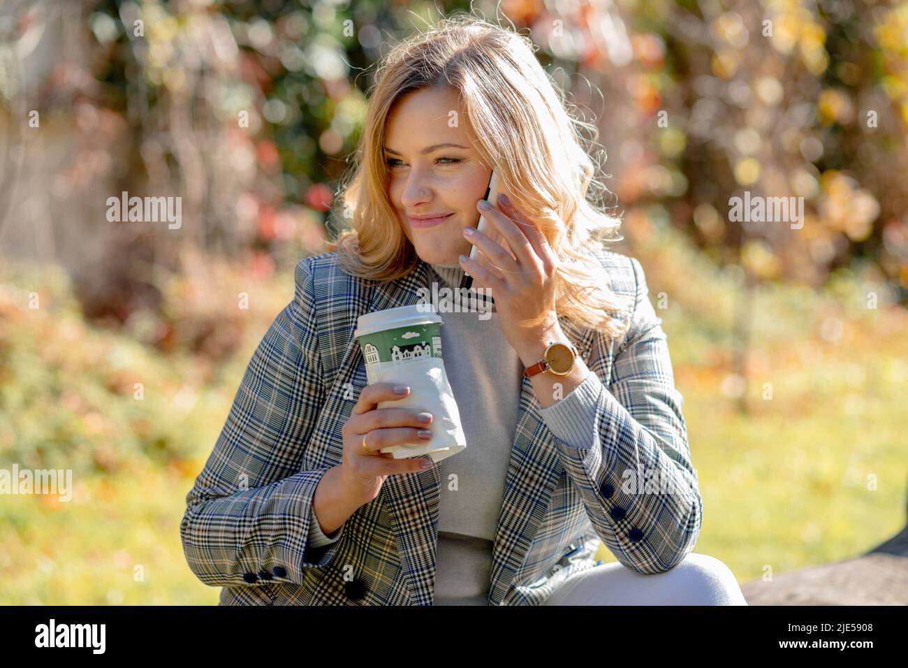 Happy young woman with long blonde hair in sweater and jacket smiles while talking on phone, sitting in city park against background of autumn trees Stock Photo