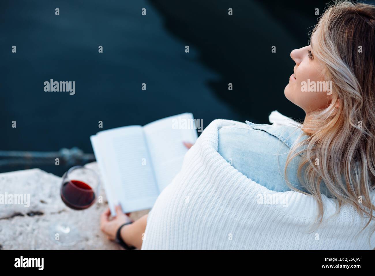 beautiful young woman is sitting on pier with glass of red wine and book having great time on vacation , happy evening. Stock Photo