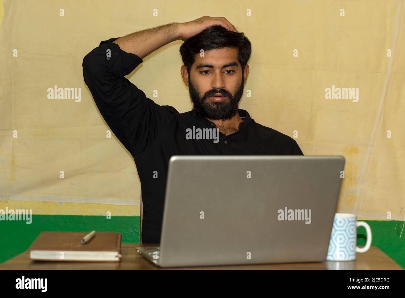 A bearded Indian man wearing black formal shirt, sitting on a chair and giving shocked expression looking at laptop screen. Diary and coffee mug on ta Stock Photo