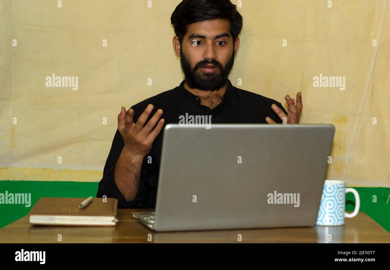 A bearded Indian man wearing black formal shirt, sitting on a chair and giving surprised expression looking at laptop screen. Diary and coffee mug on Stock Photo
