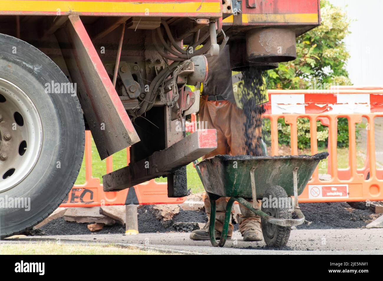A workman waits by a wheel barrow whilst its filled with fresh tarmac from a lorry. The tarmac is for repairs to a street that has been dug up Stock Photo