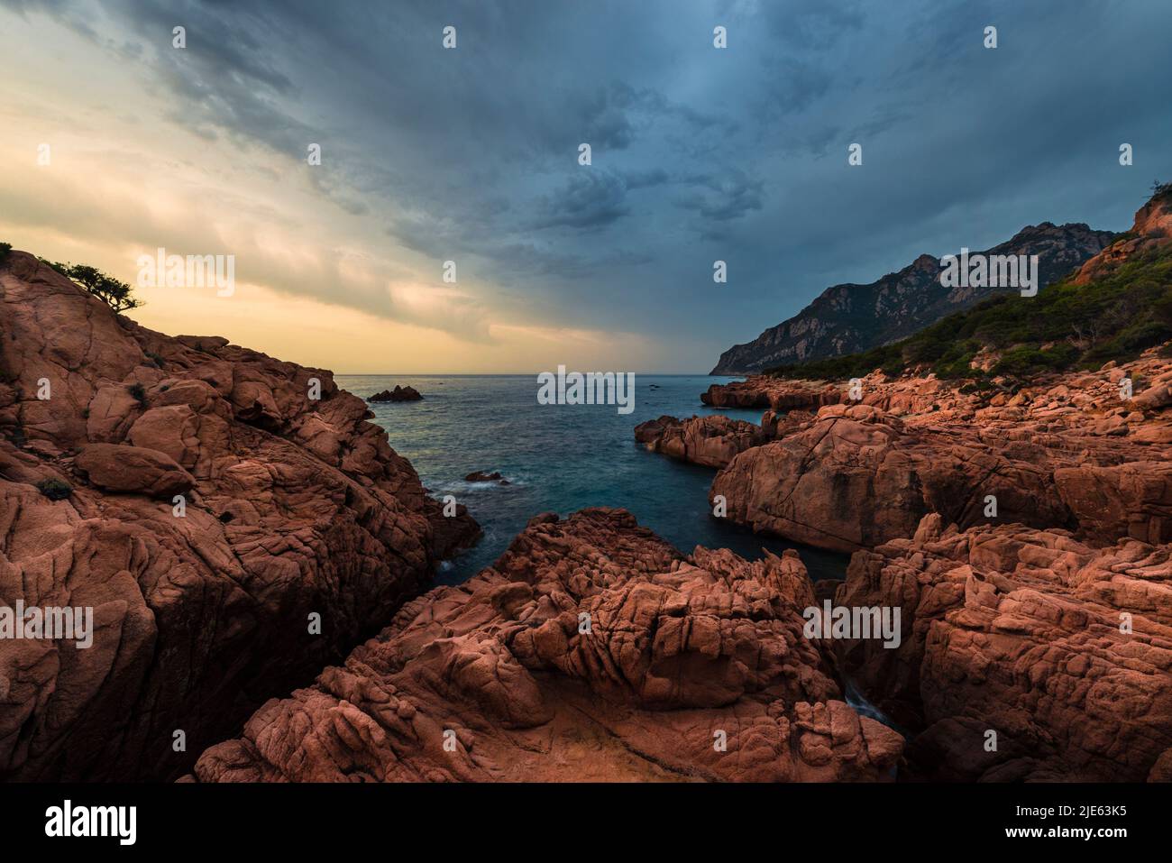Sunrise with dramatic sky at the bay of Coccorocci with the red porphyry rocks, grey pebbles and Monte Cartucceddu on the east coast of Sardinia Stock Photo