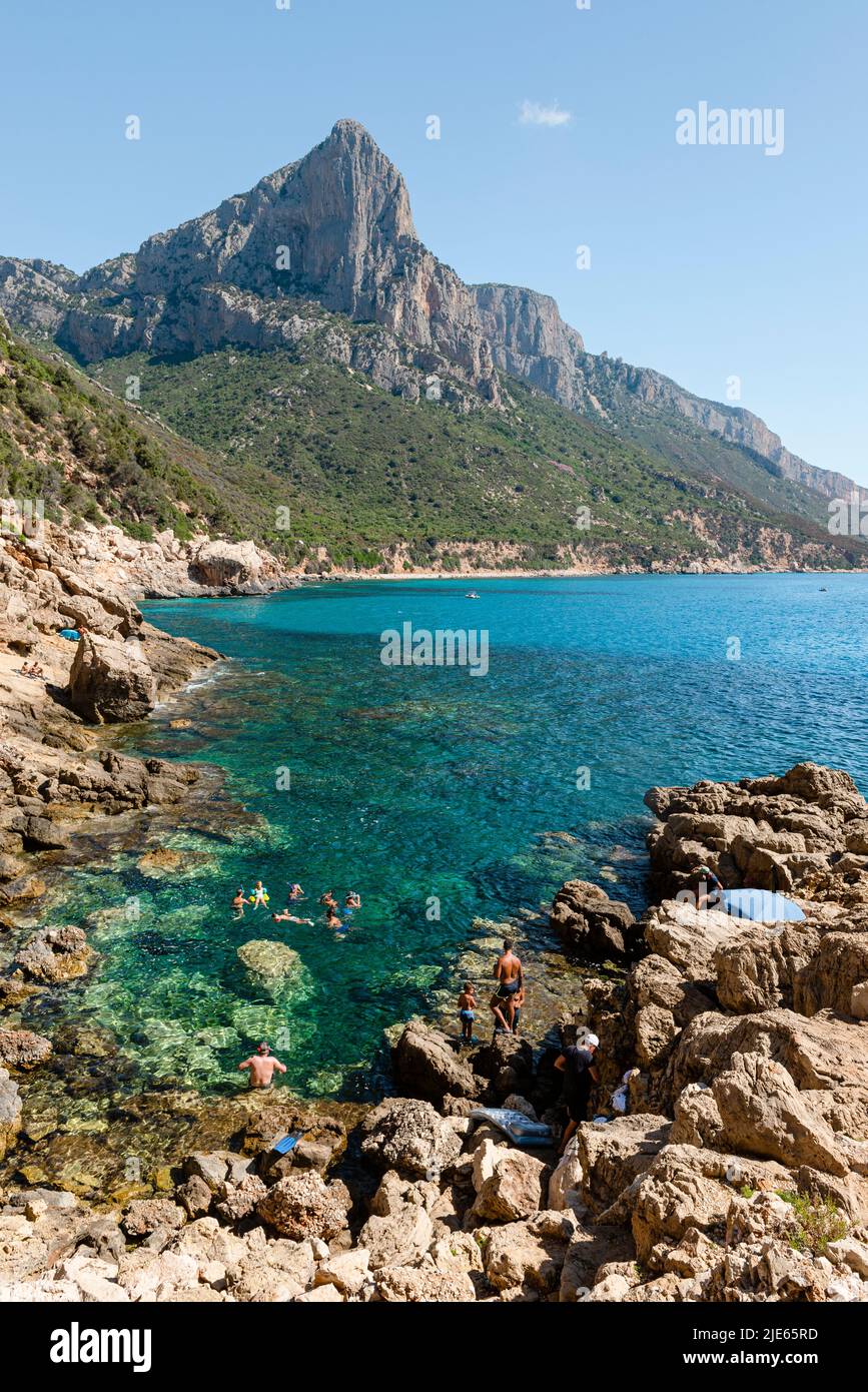 Swimmers in the turquoise sea on the cliff below the rocky walls of Punta Giradili on the east coast of Sardinia, shining in the morning sun Stock Photo