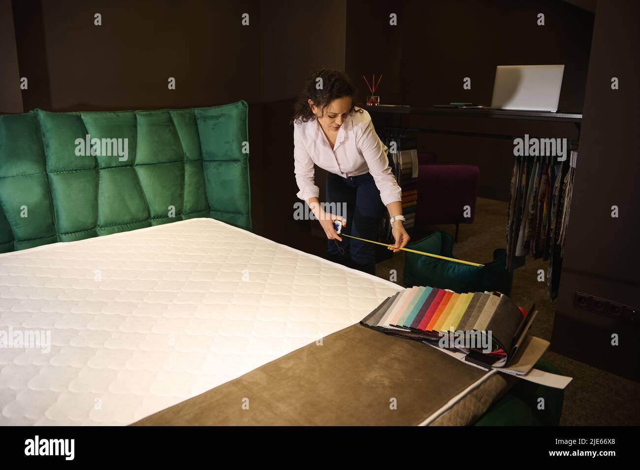 Interior designer using measuring tape measures the length of the mattress and the soft bed to order new upholstery. Upholstery fabric samples lie on Stock Photo