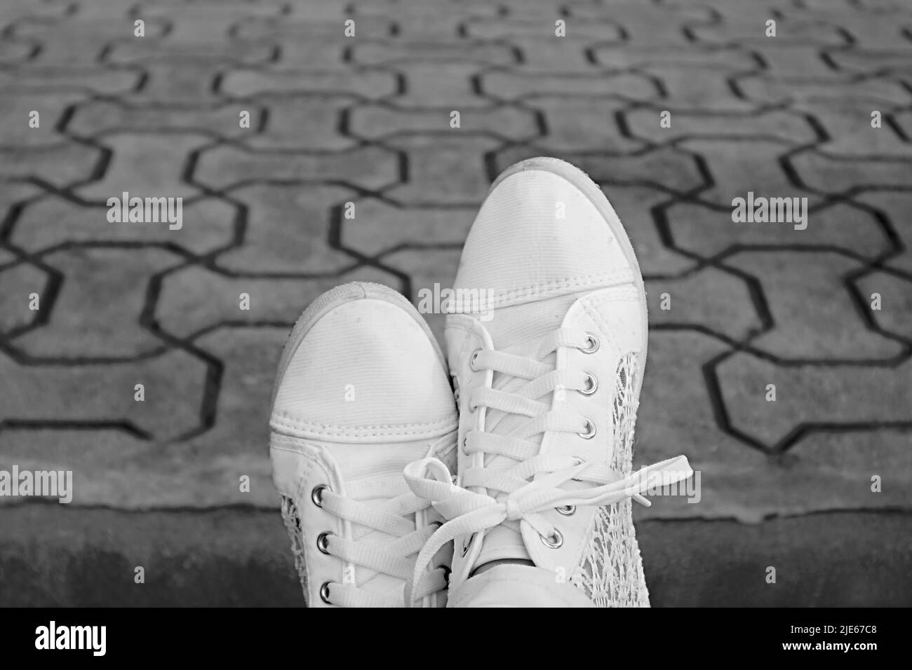 Monochrome image of feet in white sneakers relaxing on the garden's concrete block pavement Stock Photo