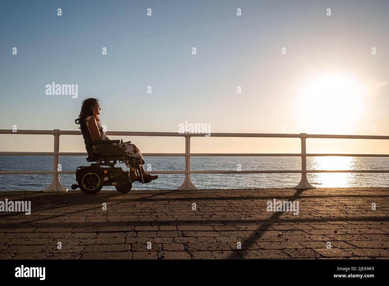 Attractive woman in an electric wheelchair gazing out to sea at sunset ...