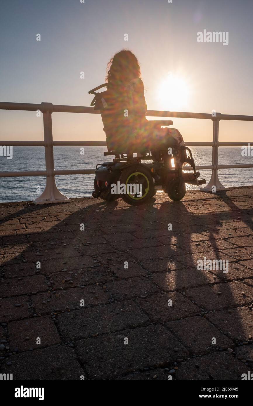 Attractive woman in an electric wheelchair gazing out to sea at sunset ...