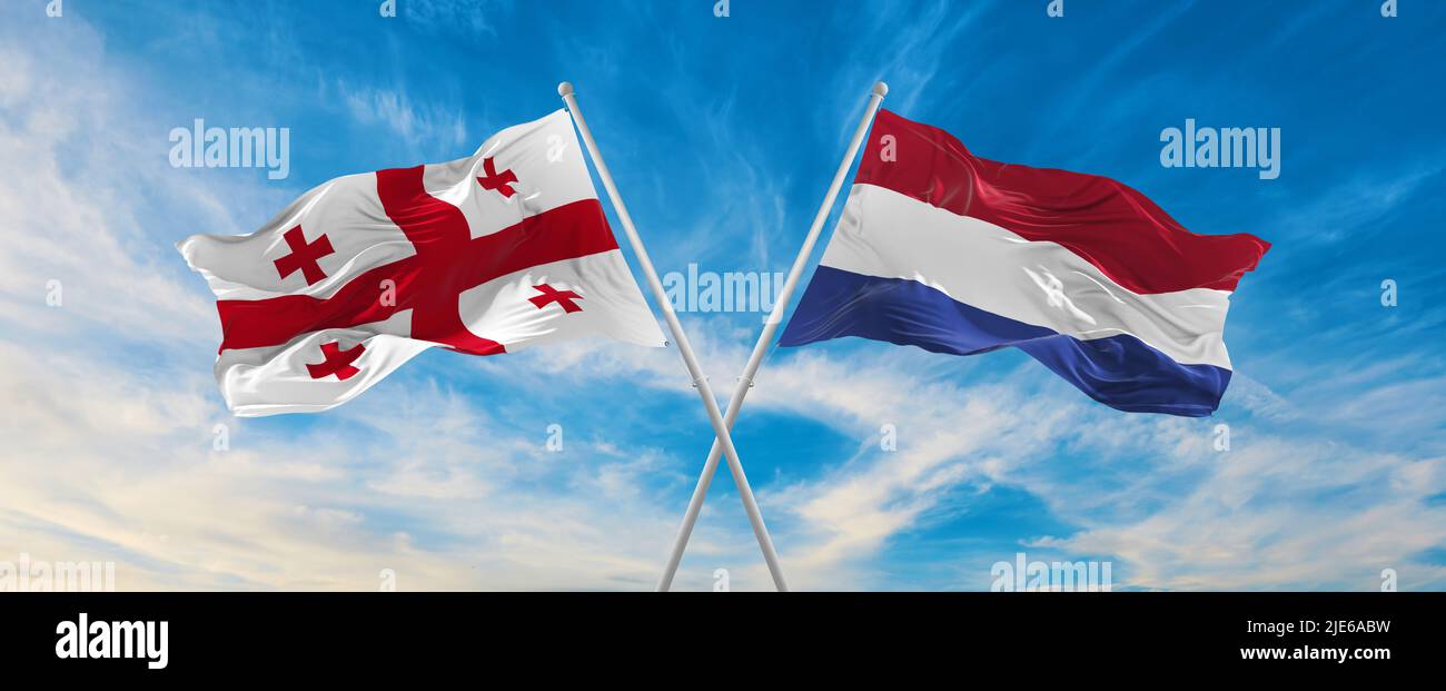 two crossed flags Georgia and Netherland waving in wind at cloudy sky ...