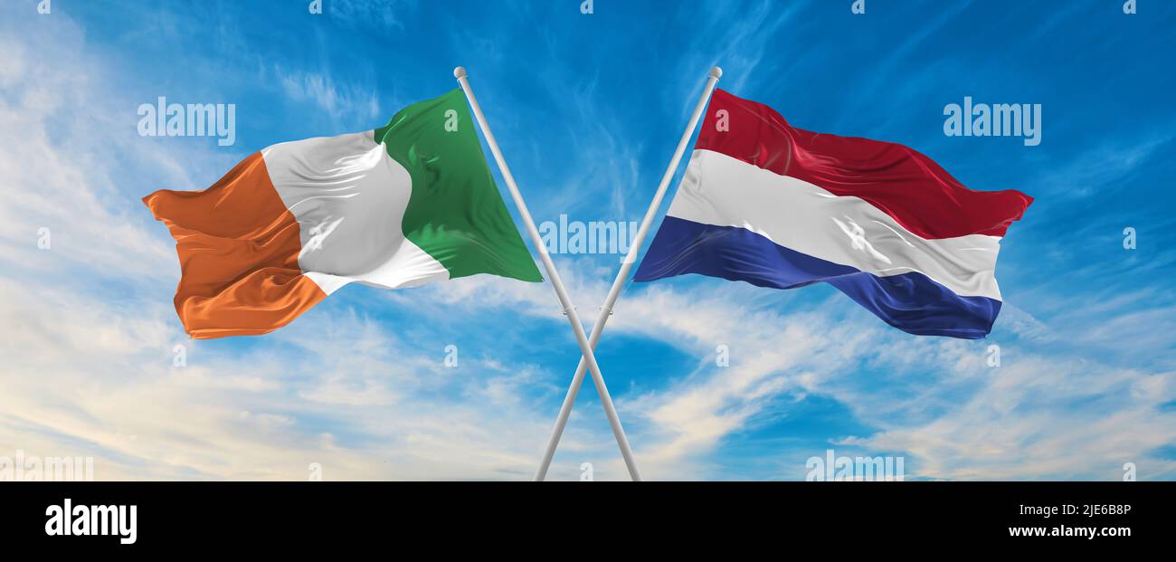 two crossed flags Ireland and Netherland waving in wind at cloudy sky ...