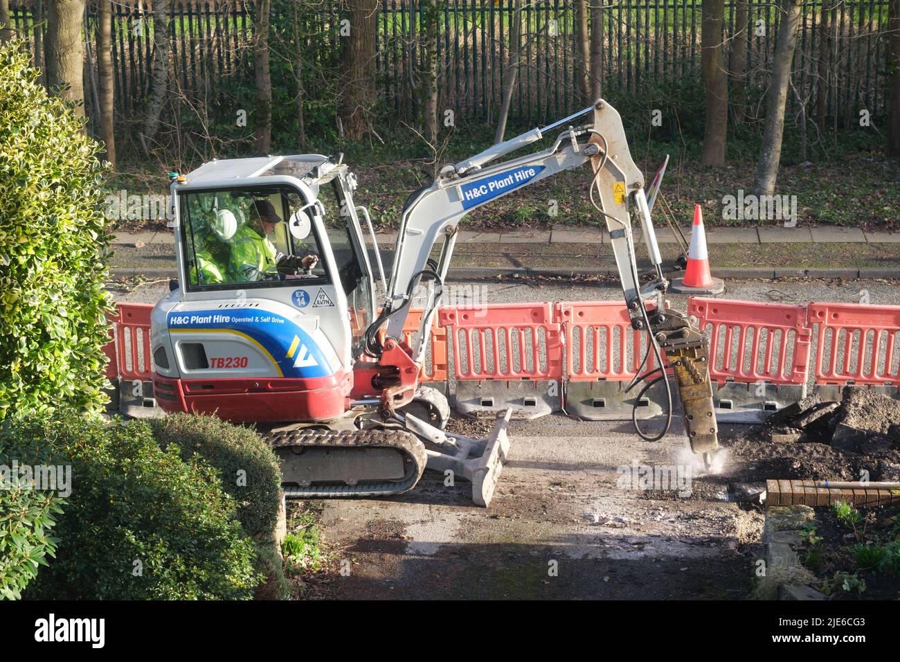 Workman using a mini excavator to dig up a pavement (sidewalk) by a road in a residential area Stock Photo